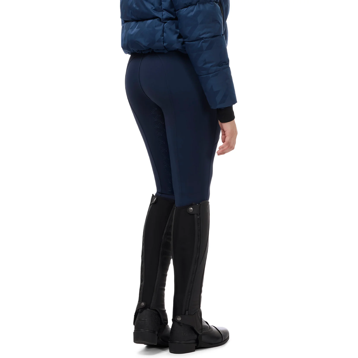 Rear three-quarter view of a model wearing nightfall colored Kerrits Fusion II Full Leg Equestrian Clinic Breeches, a navy quilted puffer jacket, and black equestrian riding boots against a white background.