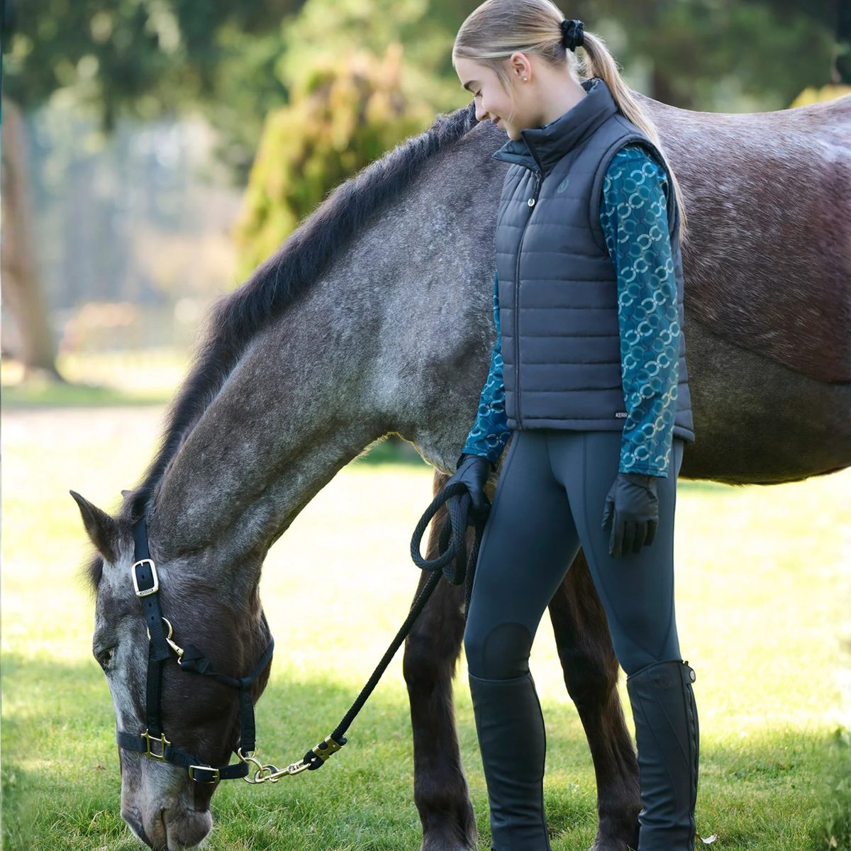 A young rider wearing Kerrits Kids Fleece Lite II Knee Patch riding tights standing  beside a dapple gray horse in a grassy outdoor setting. 
