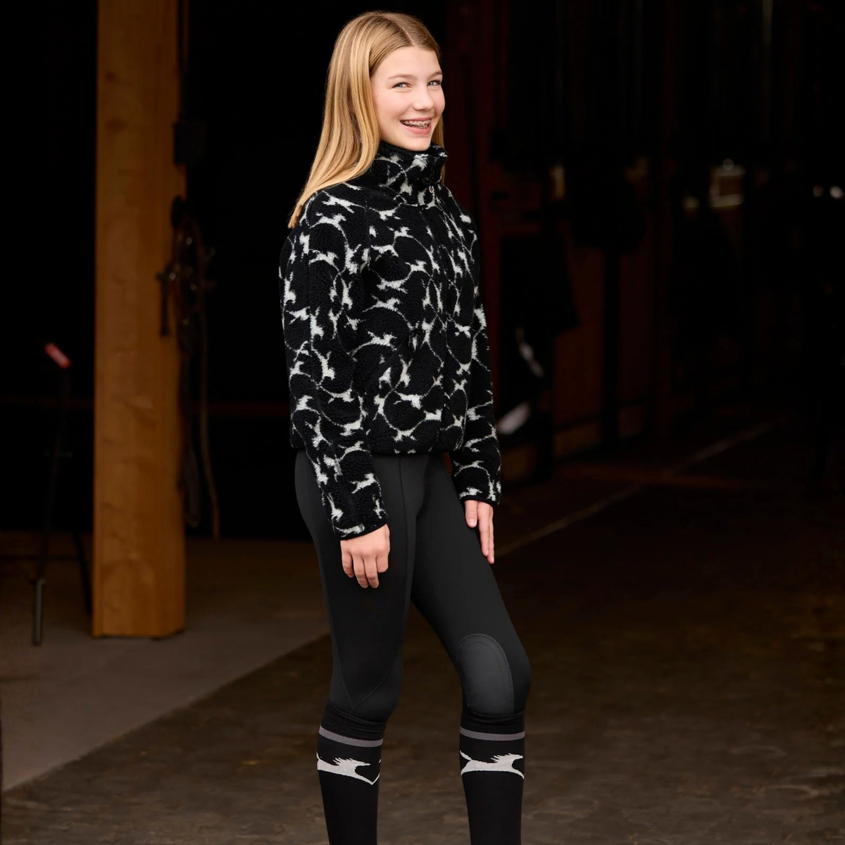 A young female rider stands in a dark  stable doorway wearing a black and white patterned fleece sweater, black Kerrits Kids Fleece Lite II Knee Patch riding breeches, and black riding boots with white stripes.