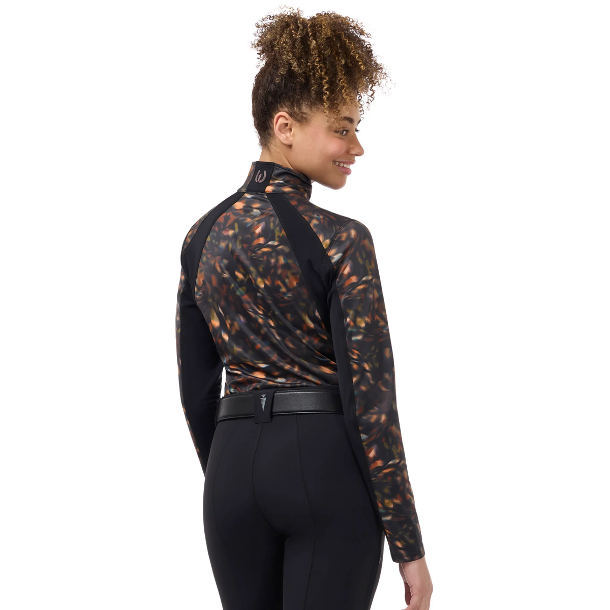 A woman shown from the back wearing a long-sleeve Kerrits Traverse Autumn Prism quarter zip fleece tech top with an abstract fall leaf patter and solid black ribbed sides paired with beige riding pants.