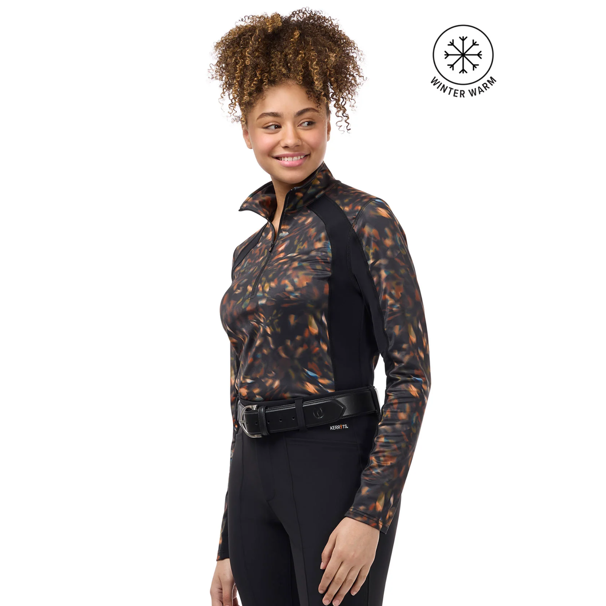 A woman modeling a long-sleeve Kerrits Traverse Autumn Prism quarter zip fleece tech top with an abstract fall leaf patter and solid black ribbed sides paired with beige riding pants. A &
