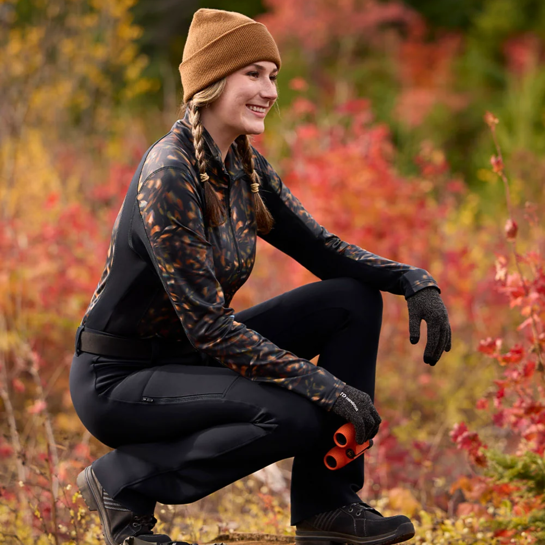 A woman crouches outdoors in a wooded area with autumn foliage, wearing Kerrits traverse quarter zip fleece tech top in Autumn Prism, black pants, black gloves, and a tan beanie. 