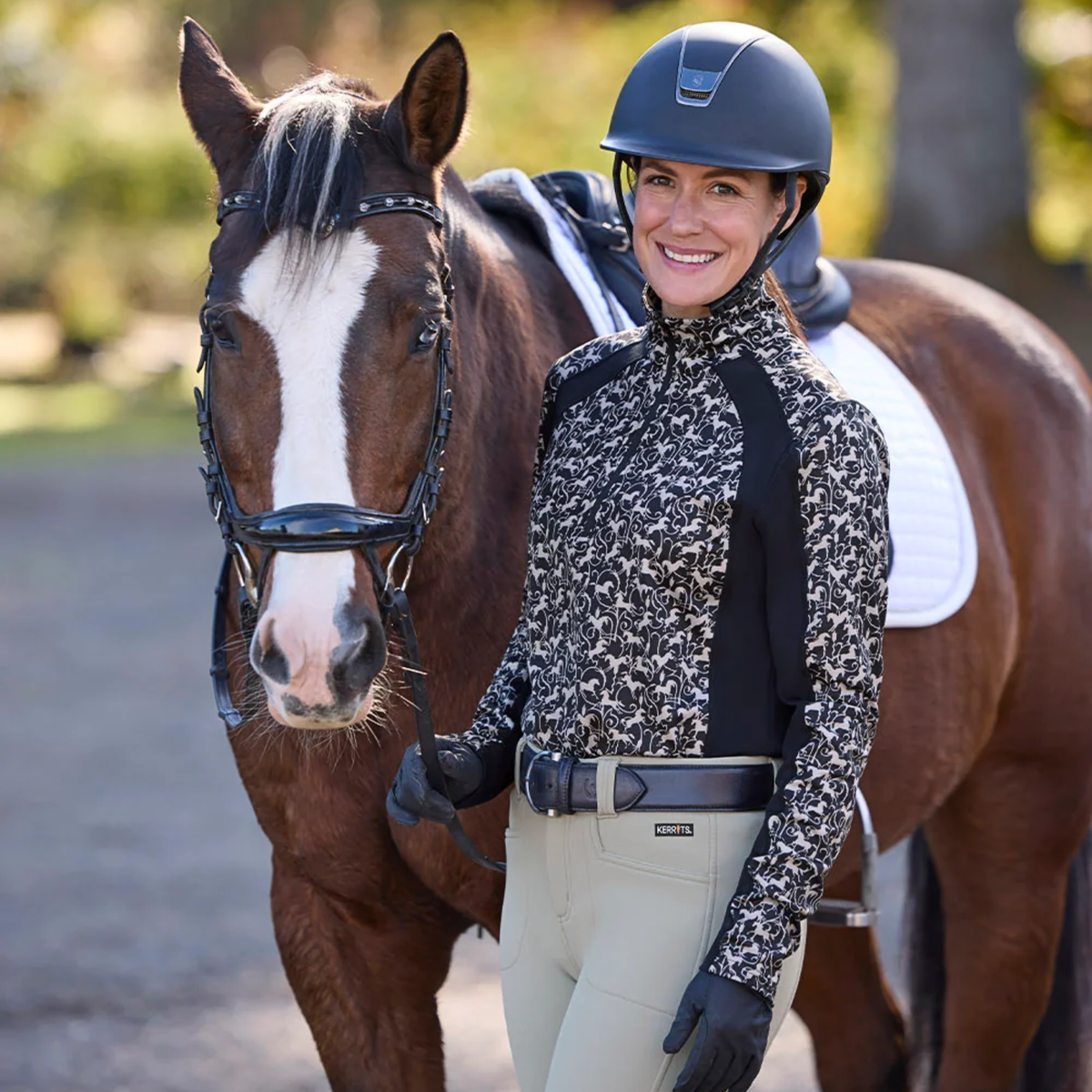A woman in equestrian apparel stands beside a brown horse wearing a black riding helmet, a  Kerrits Traverse quarter zip tech top in black wrought iron horses print, gloves, and light beige riding pants.