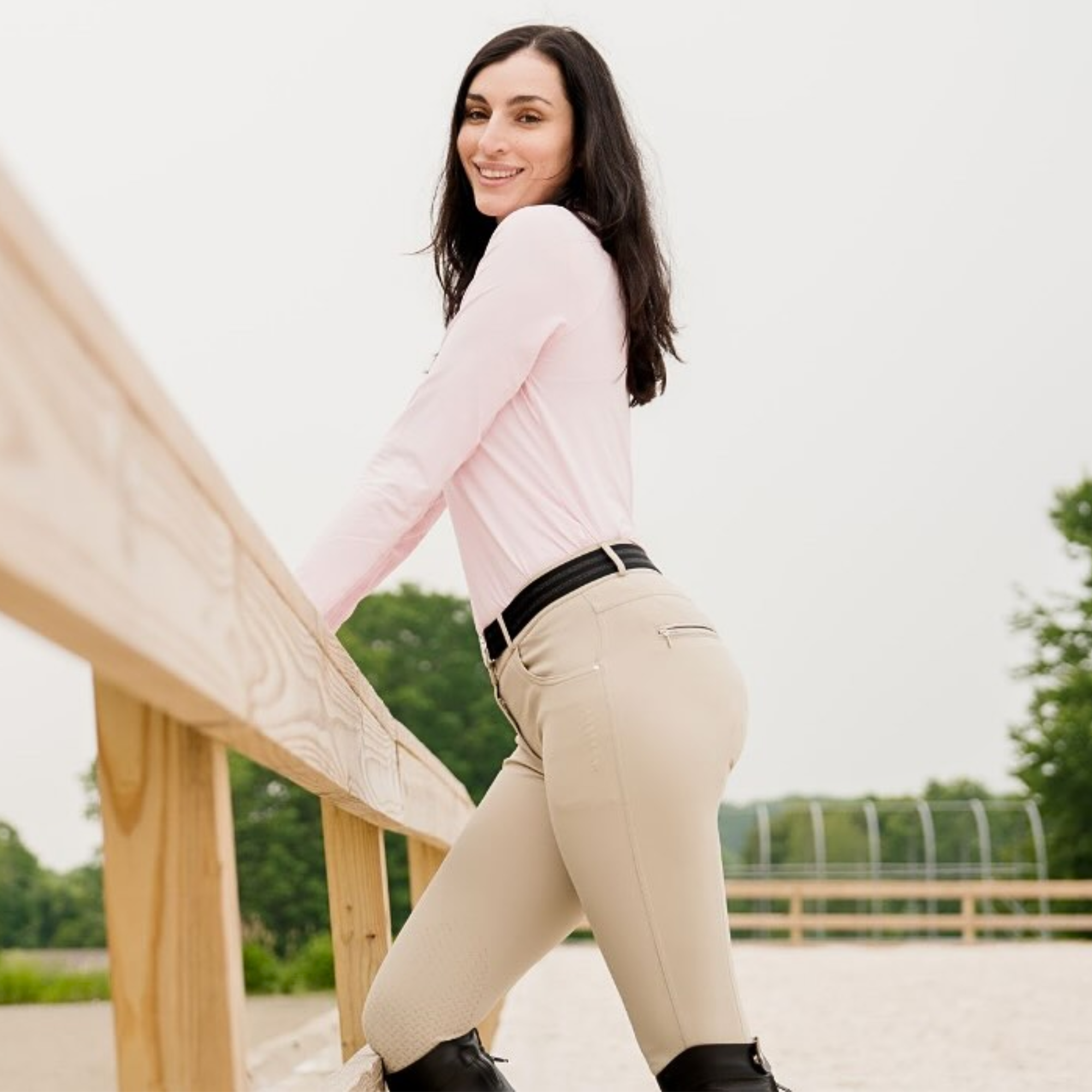 Woman in pink shirt and beige pants standing outdoors with a wooden structure and greenery in the background