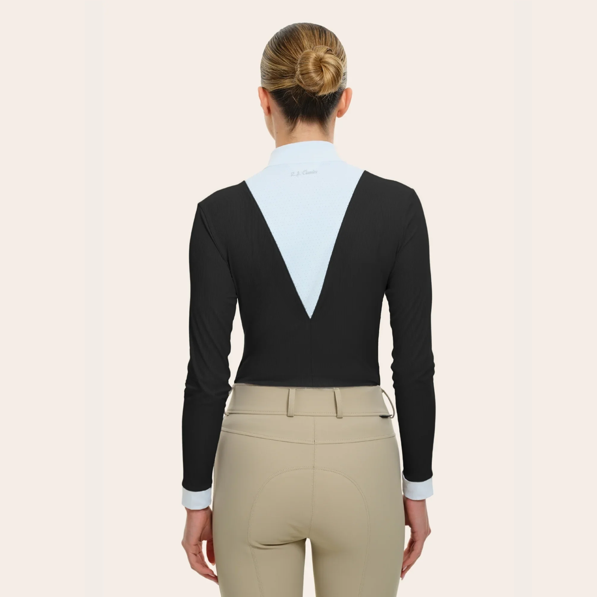 A woman viewed from the back wearing a long sleeve, black R.J. Classics Celia show shirt with a white V-shaped back panel and cuffs and beige riding breeches against a plain light beige studio background.