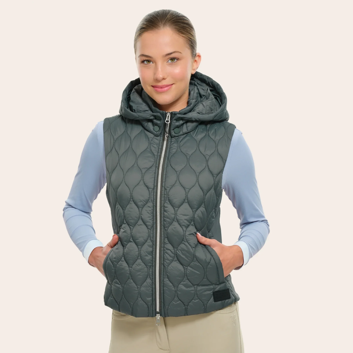 A woman wearing a dark gray/green quilted R.J. Classic Riley vest over a light blue long-sleeve shirt and beige pants, standing against a solid light beige background.