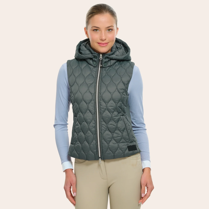A woman modeling a dark gray quilted hooded puffer vest over a light blue long-sleeve shirt and beige pants, set against a solid light beige background.