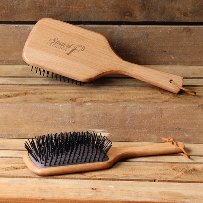 Two wooden backed, black bristled Smart Grooming Large mane &amp; tail brushes are displayed on a rustic wooden plank surface. The top brush is shown from the back, featuring a branded handle with the brand name &