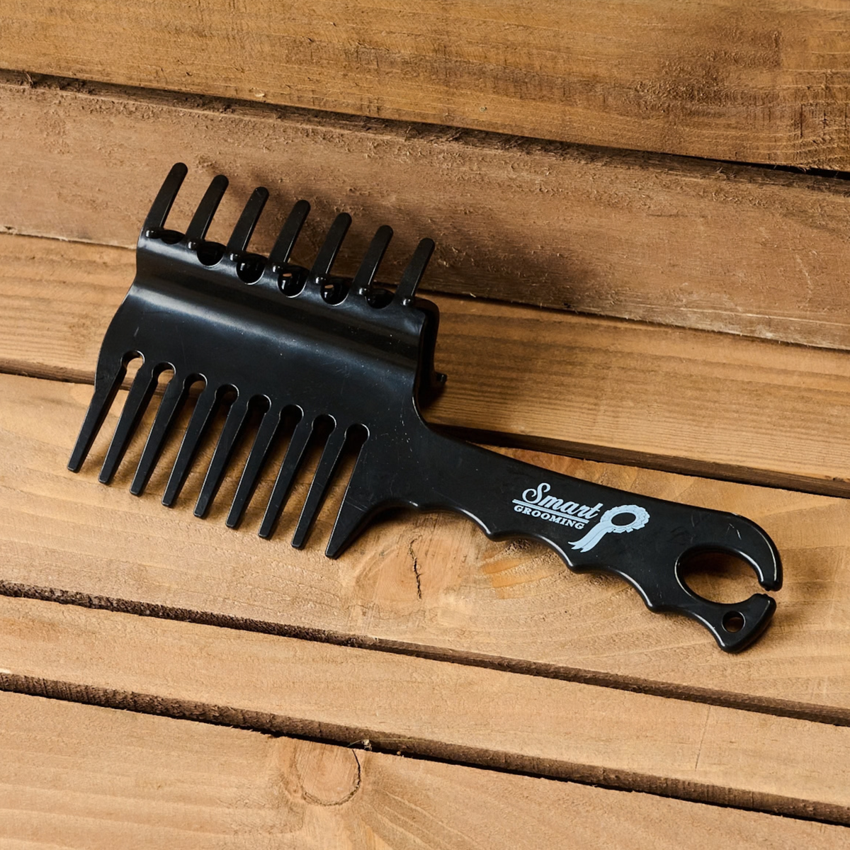 A black Smart Grooming Perfect Plaits wide-tooth comb with a shaped handle and and a built in band holder at the end, resting on a rustic wooden plank surface.