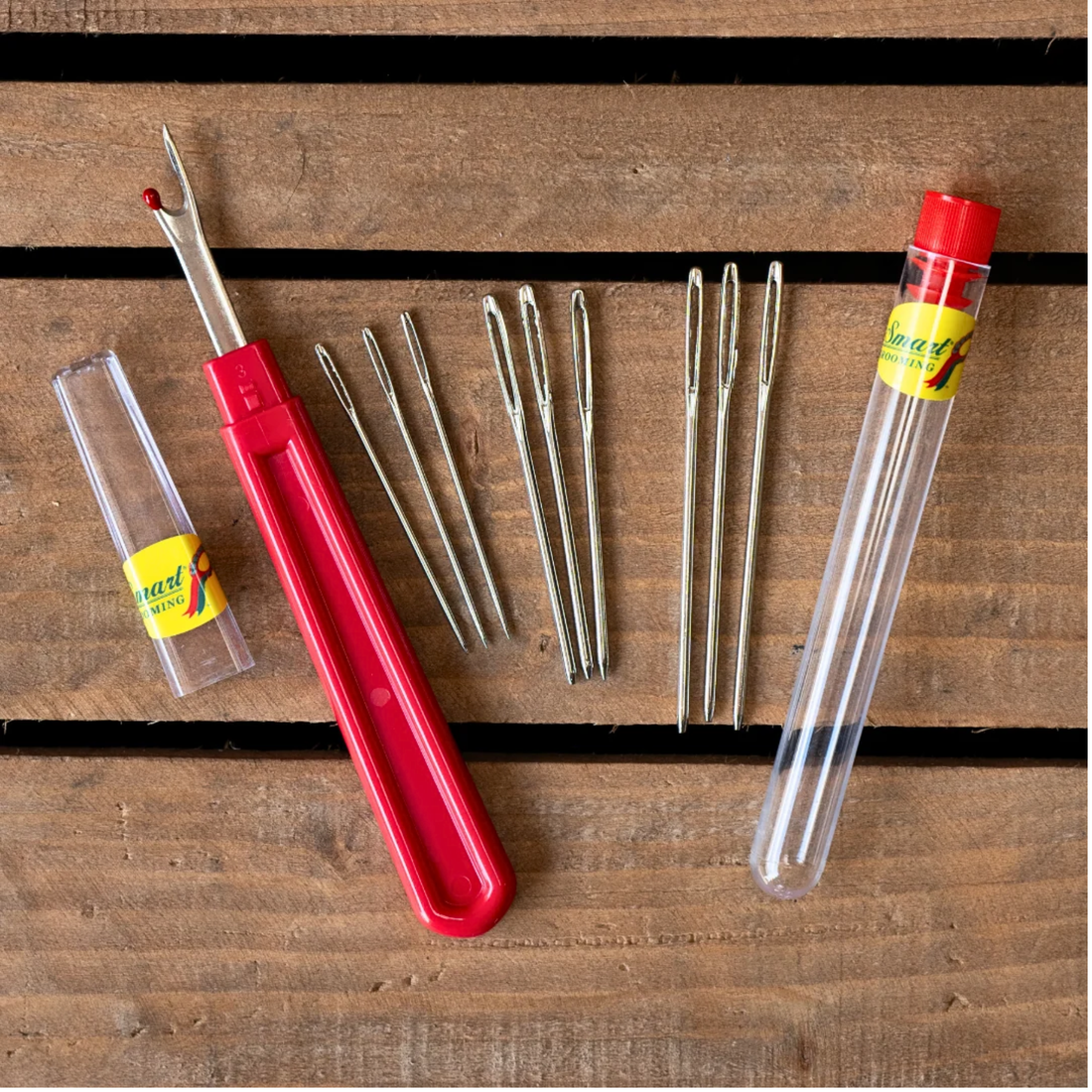 Smart Grooming braiding needs in 3 sizes  with plastic storage tube and red handled seam ripper with a clear cap laid out on a wooden surface.