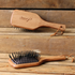 Two wooden backed, black bristled Smart Grooming small mane & tail brushes are displayed on a rustic wooden plank surface. The top brush is shown from the back, featuring a branded handle with the brand name &