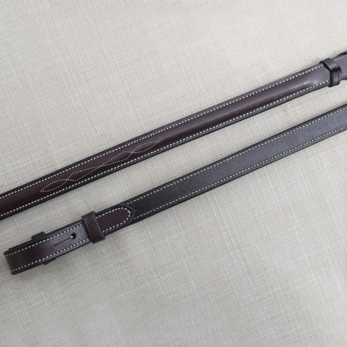 A pair of brown kl select round raised fancy lace reins with white stitching, laid out on a textured background.