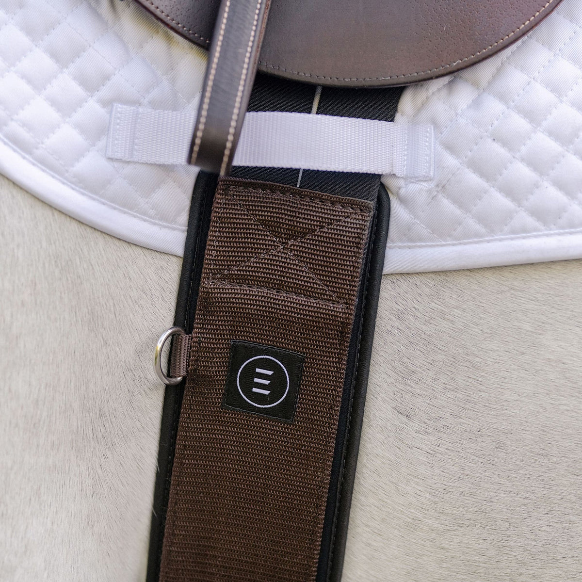 A brown EquiFit Essential Schooling Girth with a SmartFabric Liner, attached to a saddle on a white horse.