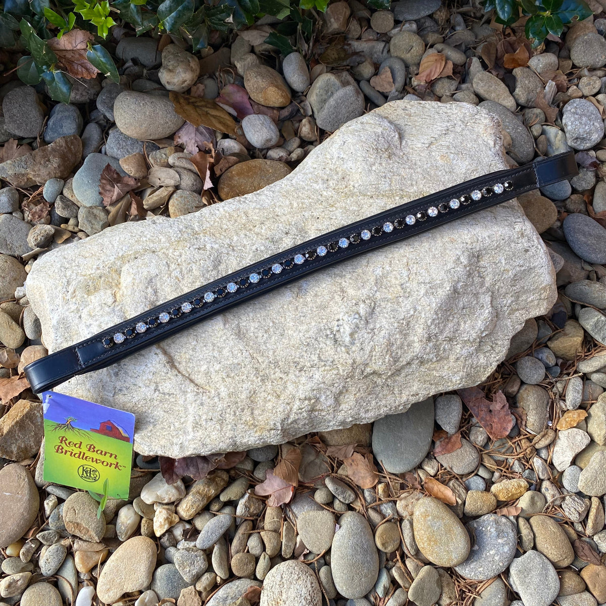 A Red Barn Bridlework black padded browband with clear crystal inserts, laid out on a rocky surface with leaves scattered around.
