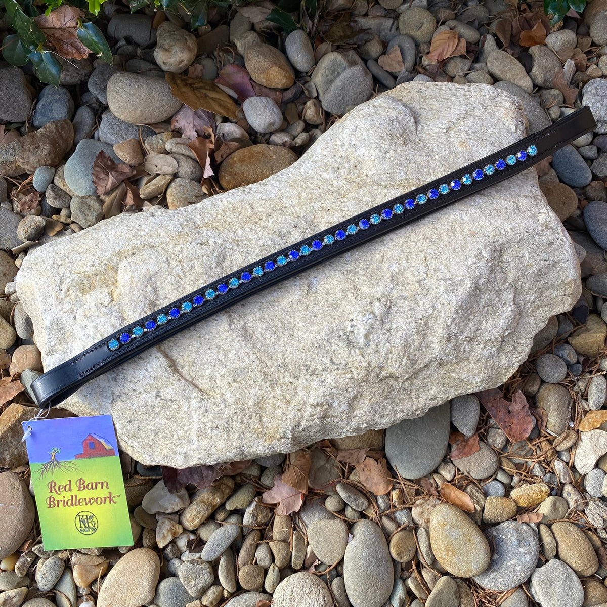 A Red Barn padded browband with large aqua and blue crystals, displayed on a rocky surface with a tag showing the brand &